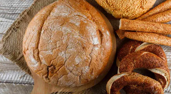 Różne rodzaje pieczywa: duża bochenek, paluszki sezamowe, obwarzanki i chleb z ziarnami.