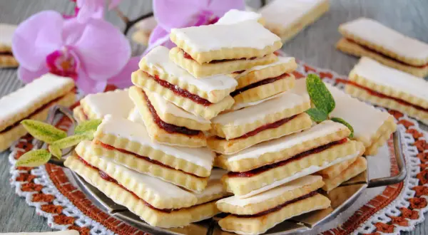 Stos ciastek przekładanych dżemem, polanych lukrem, na ozdobnej serwetce, z kwiatami w tle.